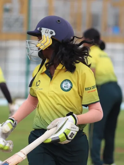 A female cricketer walking off the field, fully equipped, after a practice session or match.