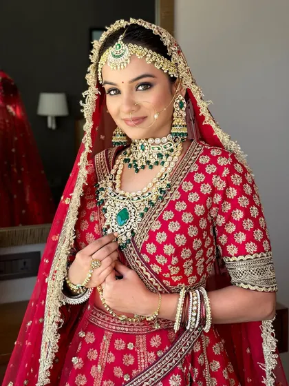 This bride looks stunning in her red lehenga, with makeup that enhances her features and complements her emerald jewelry. A perfect example of a classic bridal look.