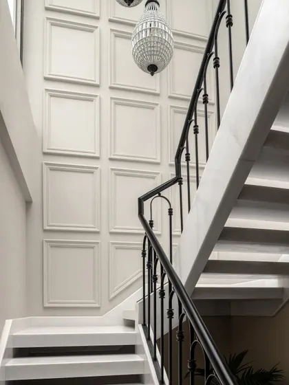 The staircase in the Quiet Ornate home features a wall of classic moulding and a stunning crystal chandelier. These details add a sense of ornate, baroque-inspired elegance to the space.