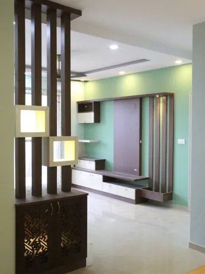 This living room features a beautiful wooden partition that separates the seating area from the pooja space. The partition includes illuminated niches for displaying decor.