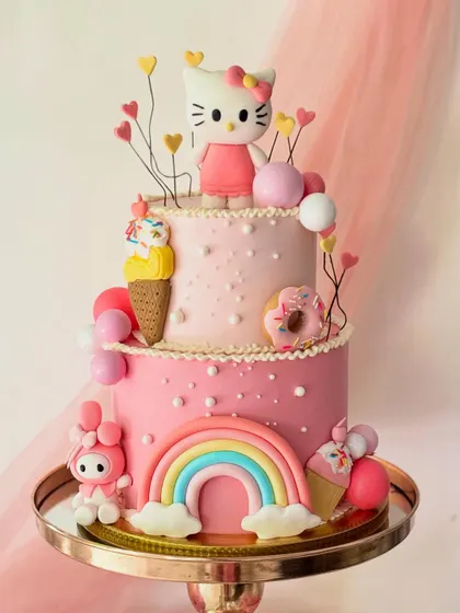 A whimsical two-tier Hello Kitty cake designed for a magical birthday. The cake is adorned with edible rainbows, ice cream cones, donuts, and beloved Sanrio characters, all crafted from buttercream and sugar paste.