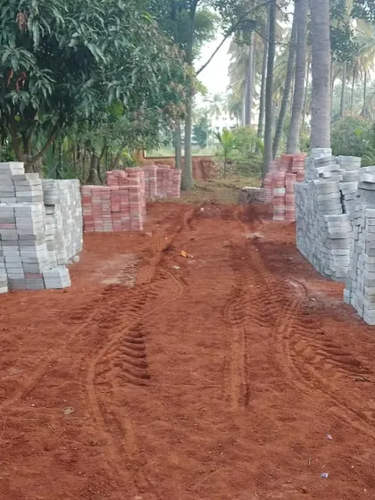 The starting point of a large farm garden project. The image shows a dirt path with stacks of paving bricks ready for installation, surrounded by mature coconut palms.