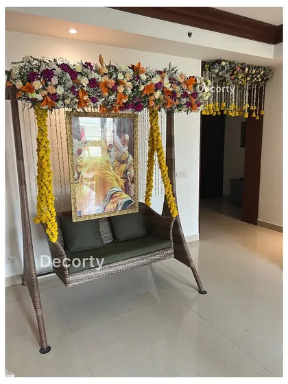 A beautifully decorated swing (jhoola) for a Mundan ceremony, adorned with yellow floral strings and a lush overhead flower arrangement. A portrait of a deity hangs in the background, adding a spiritual touch.
