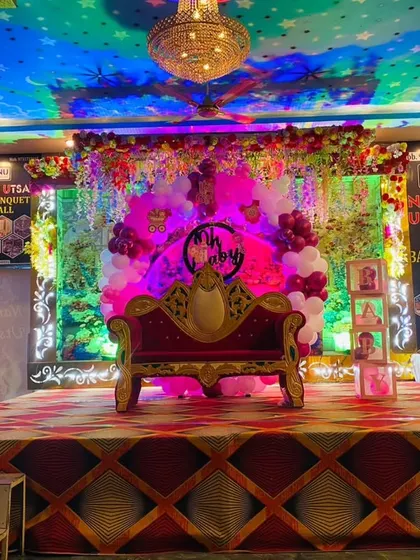 A grand stage setup for a naming ceremony in a banquet hall. The decor includes a royal-style sofa, Boss Baby character cutouts, and pink and white balloon pillars, creating a special spot for the main event.