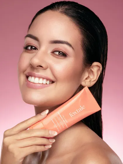 A bright, smiling portrait of a model holding a tube of Foxtale's SPF primer. Her happy expression and flawless skin make a compelling case for the product. This is a classic commercial beauty shot that radiates positivity and confidence.