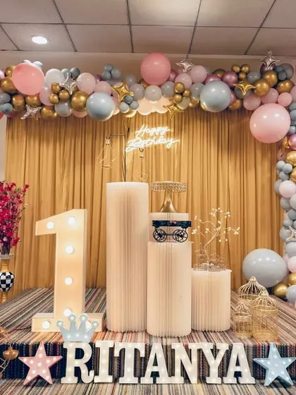We created this beautiful setup for Ritanya's first birthday party. It features a custom balloon arch in pastel pink, blue, and gold, a glowing 'Happy Birthday' neon sign, and a large light-up number '1'. The little white picket fence and decorative plinths for the cake and props added a charming touch to the entire photo corner.