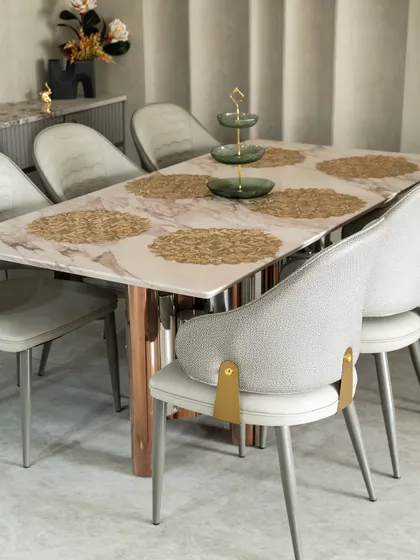 This dining setup exudes quiet luxury. The marble tabletop with soft pink veining is paired with chairs accented by brushed gold brackets. Every detail, from the golden placemats to the tiered centerpiece, is curated for a refined dining experience.