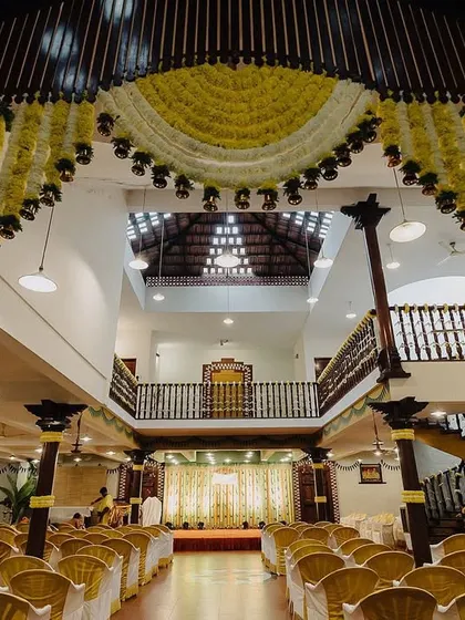 A wide shot of a beautifully decorated wedding hall. The grand floral arch and traditional seating arrangement showcase the scale and elegance of the event's decor.