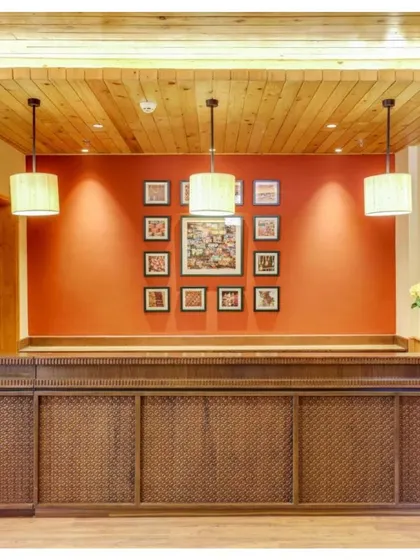 The reception desk at Janjehli Resort features intricate wooden carving and is set against a bold red wall adorned with traditional Chamba Rumal artwork. This immediately immerses guests in the local culture.