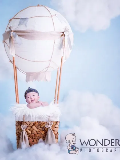 Up, up, and away! A happy baby smiles from a hot air balloon basket prop, floating among fluffy clouds against a bright blue sky. This is one of my most popular and imaginative concepts.
