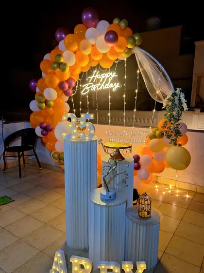 A vibrant rooftop birthday surprise with a circular balloon arch in orange and purple. The setup includes a custom 'Happy Birthday' neon sign and tiered pedestals for the cake and decor.