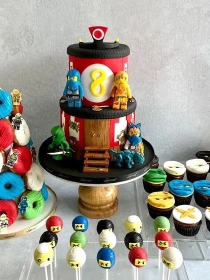A full Ninjago dessert table for Ved's 8th birthday, complete with a two-tiered cake, a donut tower, and matching cupcakes and cake pops.