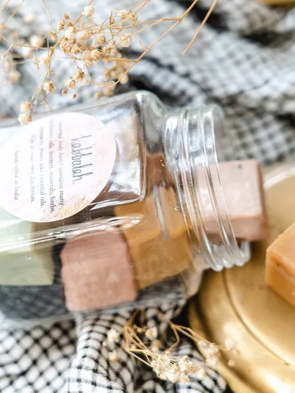 A jar of my assorted soap cubes spilling onto a brass plate. It shows the variety of natural colors, from the deep black of charcoal to the soft pink of rose clay.