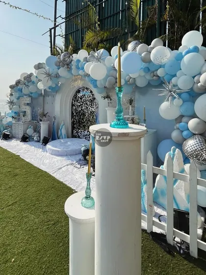 An absolutely stunning Frozen-themed setup by the sea. The backdrop features a massive balloon arch in shades of blue, white, and silver, with a sparkling sequin door.