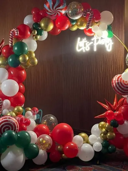 A classic Christmas party ring backdrop. The arch is filled with red, green, white, and gold balloons, along with candy cane and star accents, all lit by a "Let's Party" neon sign.