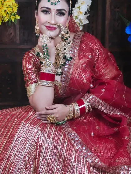 A smiling bride wearing a traditional red lehenga with a heavy green and gold Kundan jewellery set. The set includes a choker, nath, maang tikka, and haath phool.