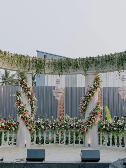 Inspired by classic Roman architecture, this outdoor stage features grand white pillars and a balustrade, entwined with lush greenery and flowers. The hanging chandeliers add a touch of timeless elegance to the structure.