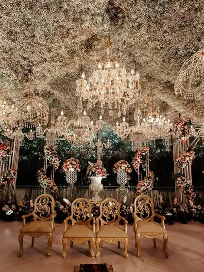 A view from within the mandap, looking out. The space is framed by elegant gold chairs for the couple and a ceiling dense with foliage and sparkling chandeliers.