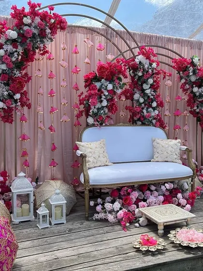 An elegant Mehendi seating area with a pink and white color scheme. The backdrop features hanging tassels and is framed by beautiful floral arches.