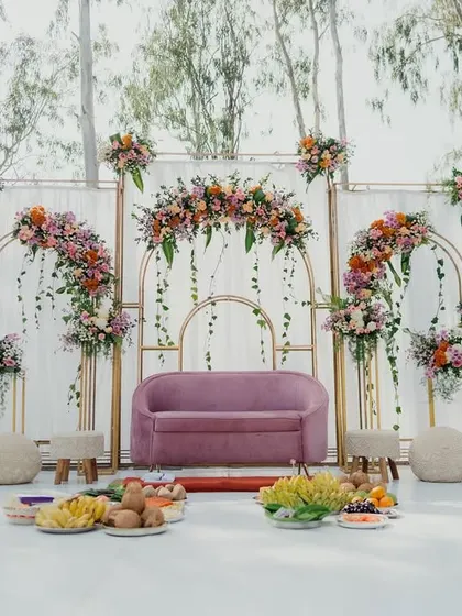 A beautiful pastel-themed stage for a daytime engagement. The setup includes elegant gold arches, a modern pink sofa, and lush floral arrangements with a mix of roses and marigolds, perfect for an outdoor celebration.