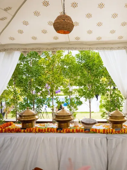 An elegant outdoor food buffet set up under a white tent. The brass serving dishes and marigold garlands add a touch of traditional Indian flair to the event.