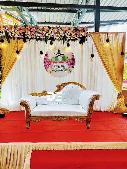 A beautiful stage setup for a 60th birthday celebration. The decor features a floral arch, elegant white and gold drapes, and a classic diwan for the honored couple.