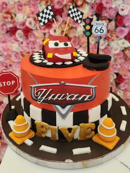 A two-tiered cake inspired by the movie 'Cars'. It features a checkered flag pattern, road signs, and Lightning McQueen on top, making it perfect for a young car enthusiast's birthday.