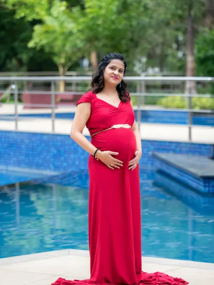 A serene solo portrait of a mom-to-be in a classic red dress, standing by a bright blue swimming pool. It's a simple yet elegant setting for a maternity shoot.