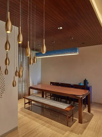 Dramatic pendant lighting creates a stunning focal point above this long wooden dining table. The minimalist setup with a bench and chairs is perfect for large family gatherings, set against a wall with a subtle, intricate pattern.