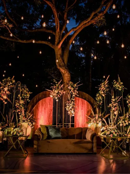 An enchanting evening setup for a wedding reception or sangeet, with fairy lights in a large tree creating a magical, starlit canopy for the celebration below.