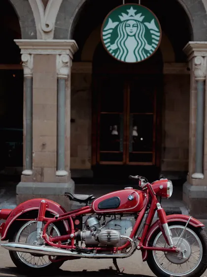 A striking vintage red BMW motorcycle is parked outside a modern Starbucks, creating a beautiful contrast between classic German engineering and contemporary culture. This urban automotive shot highlights the bike's timeless appeal in a familiar city setting.