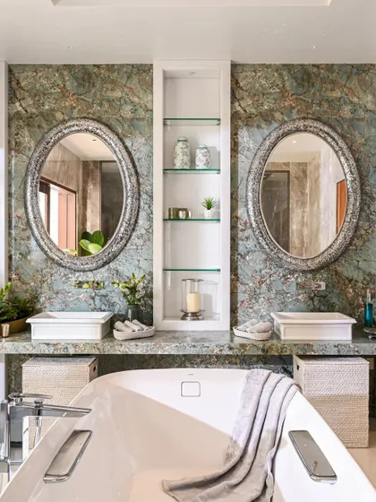 This view shows the dual vanities in the master bathroom, set against a stunning wall of green-veined marble. The matching oval mirrors with ornate frames add a touch of classic elegance to the natural stone backdrop.