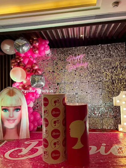 A glamorous Barbie theme for a first birthday, featuring a silver sequin wall and a neon sign. The setup includes a Barbie head cutout, themed plinths, and a life-size Barbie box photo booth for the perfect picture.