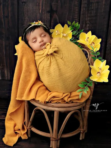 A baby in a yellow flower-style wrap, posed sitting up on a small rattan chair.