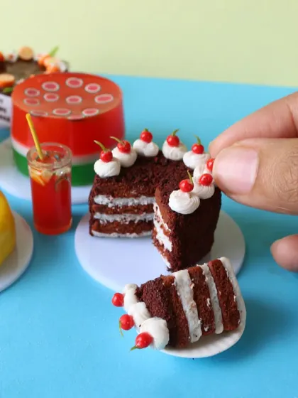 A hand holding a slice of a miniature layered red velvet cake, showing the intricate layers and frosting.