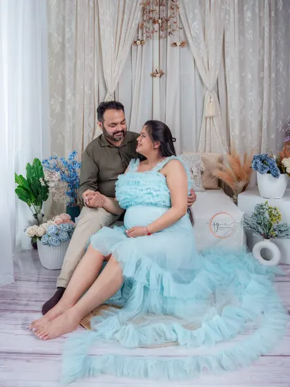 A romantic and dreamy maternity portrait. The couple is seated in a beautifully decorated boho-themed studio setup, holding hands and sharing a look of love and excitement.
