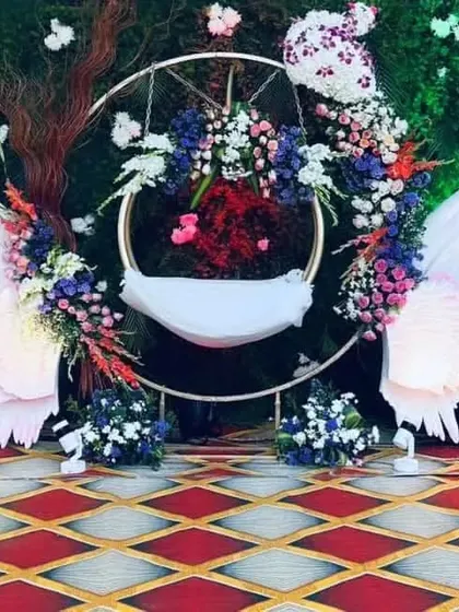 I design unique and modern cradle setups for naming ceremonies. This angel-wing themed 'thottilu' creates a heavenly centerpiece for your baby's special day, combining artistic cutouts with lush floral arrangements.