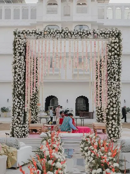 An elegant and understated mandap design, featuring a structure covered in dense white florals with delicate pink floral strings, set against the beautiful white architecture of the palace venue.