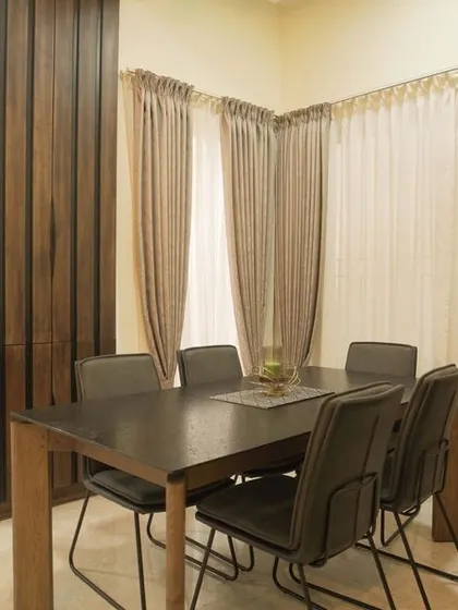 A simple yet elegant dining setup adjacent to the living area. The dark wood table and chairs are complemented by a wooden pillar and soft, neutral curtains.