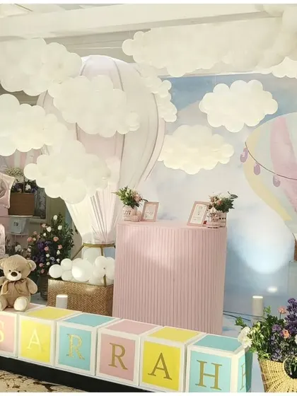 A whimsical hot air balloon themed party. The setup features a sky-blue backdrop, fluffy white clouds, and large name blocks spelling "SARRAH" in front of a pink dessert bar.