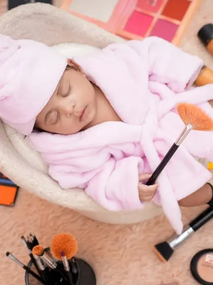 A fun and creative newborn setup featuring a baby in a pink robe and head towel, pretending to have a spa day. Surrounded by miniature makeup props, this shot is full of personality and charm.