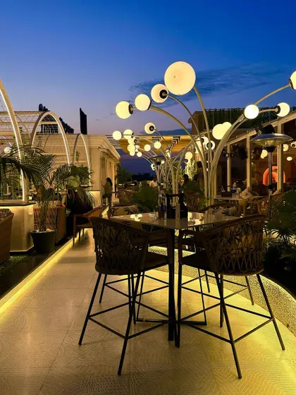 The biophilic design of the Tipsy Town rooftop at dusk. The combination of high-top seating, woven chairs, lush plants, and artistic lighting creates a strong connection with nature, offering a tranquil escape from the city.