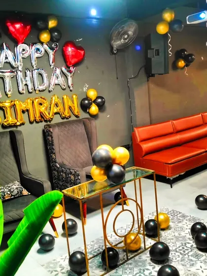 A birthday setup for Simran in a cafe, featuring black and gold balloons and "Happy Birthday" foil letters. The high-back chairs create a special seating area for the guest of honor.