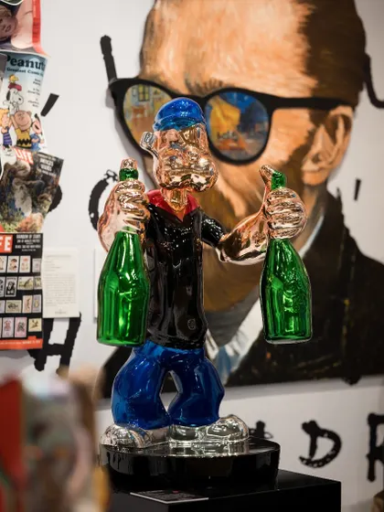 This sculpture of Popeye, rendered in metallic tones and holding green bottles, reimagines the classic cartoon character as a modern icon. It stands before a contemporary portrait, creating a layered visual effect.