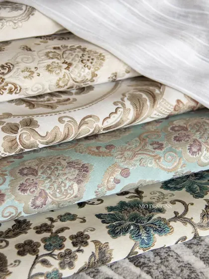 An exquisite collection of stacked fabrics, showcasing intricate floral and damask patterns in a range of colors from deep blue to soft aqua and neutral beige.