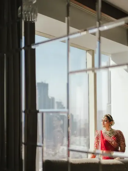 The bride looks out of a large window overlooking the city, a moment of quiet reflection before her wedding. The reflection in the mirror adds a creative, layered effect to the portrait.