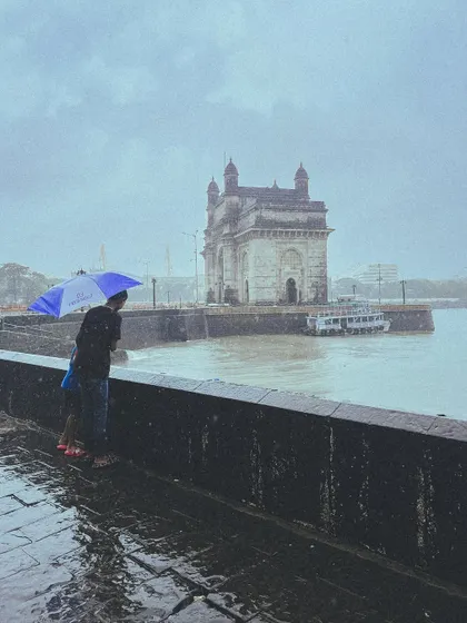 A couple stands by the sea wall, looking out at the Gateway of India, their shared umbrella a small shield against the vast, rainy expanse.