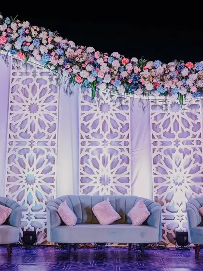 A grand and intricate stage backdrop featuring Moroccan-style geometric patterns. The arch of pastel-colored flowers and the plush blue seating create a look of sophisticated, multicultural elegance.