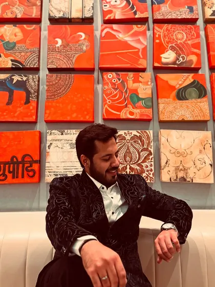 A relaxed moment in a stylish blazer, seated against a wall of traditional Indian art.