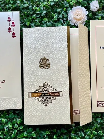 An elegant off-white invitation set with a subtle texture and gold foil accents. The design includes a Ganesha emblem and traditional bell motifs.